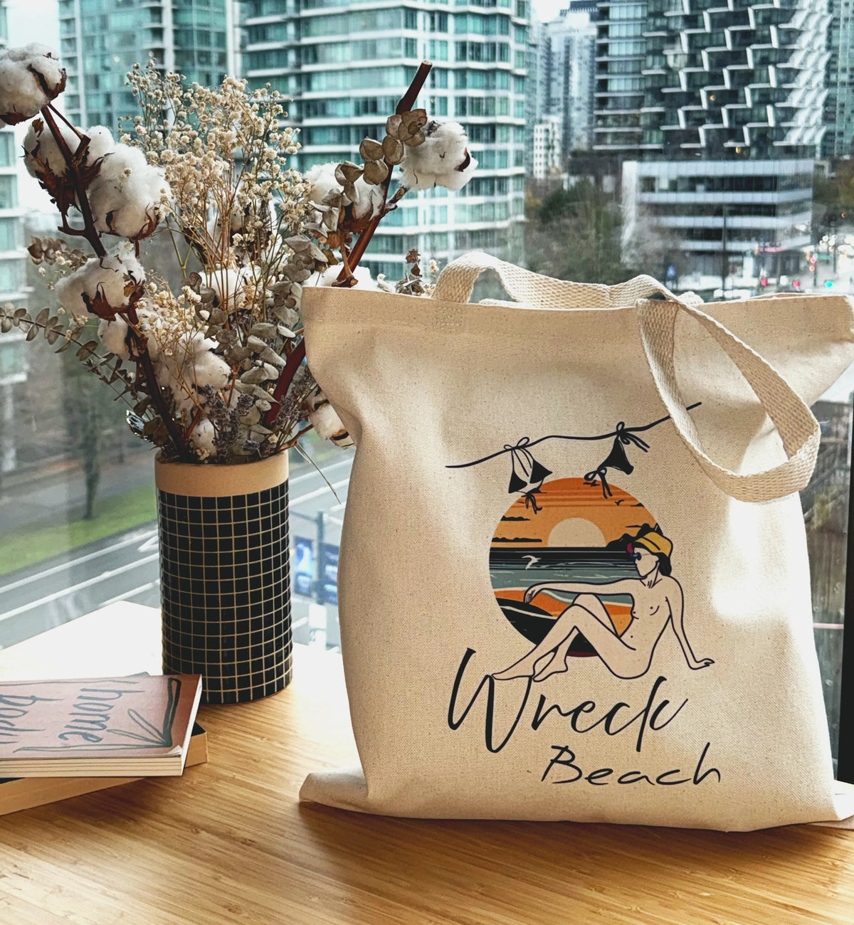 Wreck Beach canvas tote bag displayed on a wooden table beside dried flowers with city skyline in background