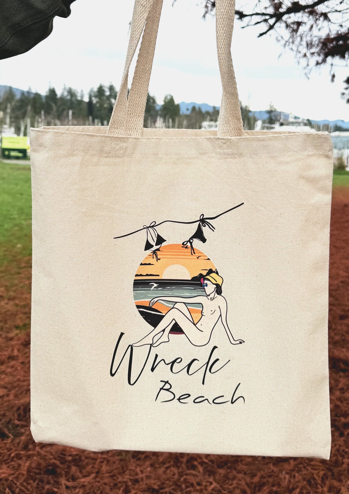 Front view of Wreck Beach canvas tote bag with sunset illustration hanging outdoors in a park