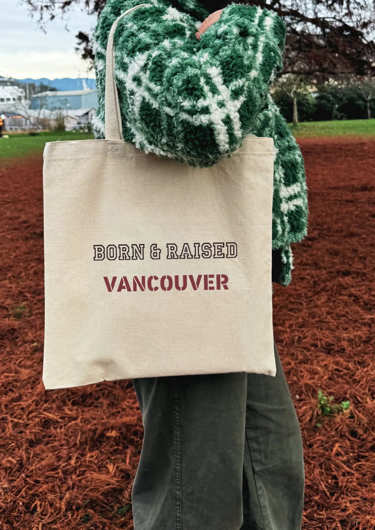 Person holding a beige tote bag with 'Born & Raised Vancouver' text, wearing a green patterned jacket outdoors.
