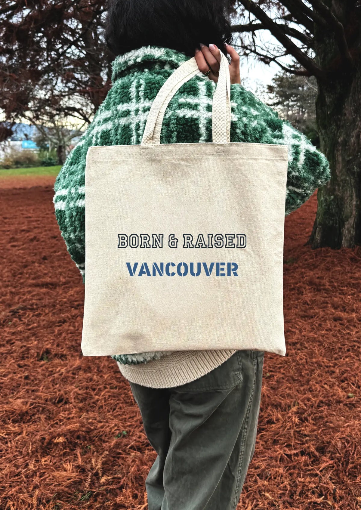 Person holding a tote bag with 'Born & Raised Vancouver' text in an outdoor setting.