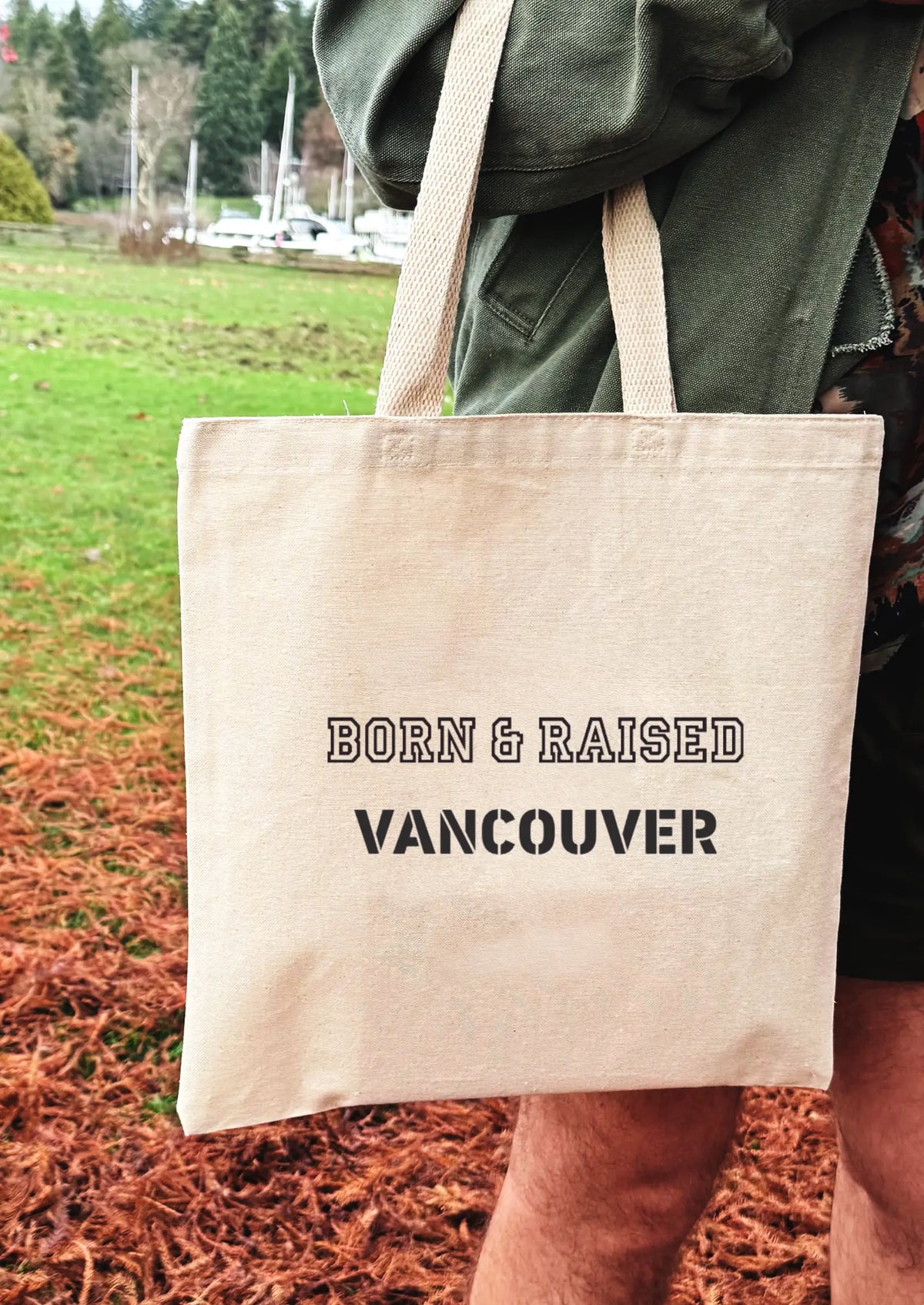 Tote bag with 'Born & Raised Vancouver' text held by a person outdoors.