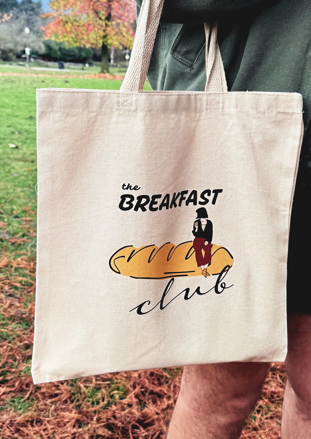 Breakfast Club canvas tote bag worn over the shoulder, featuring line art bread illustration, photographed outdoors.