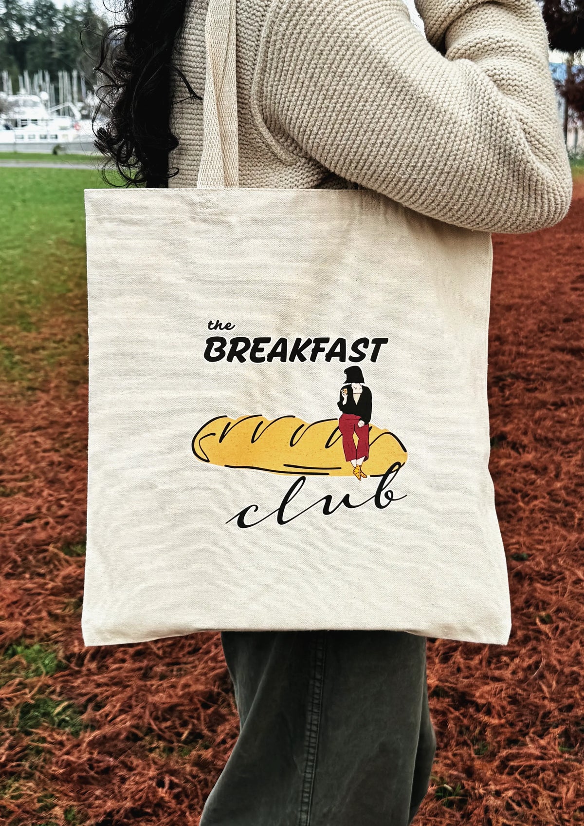 Breakfast Club canvas tote bag with bread illustration, held by hand outdoors in a park setting.