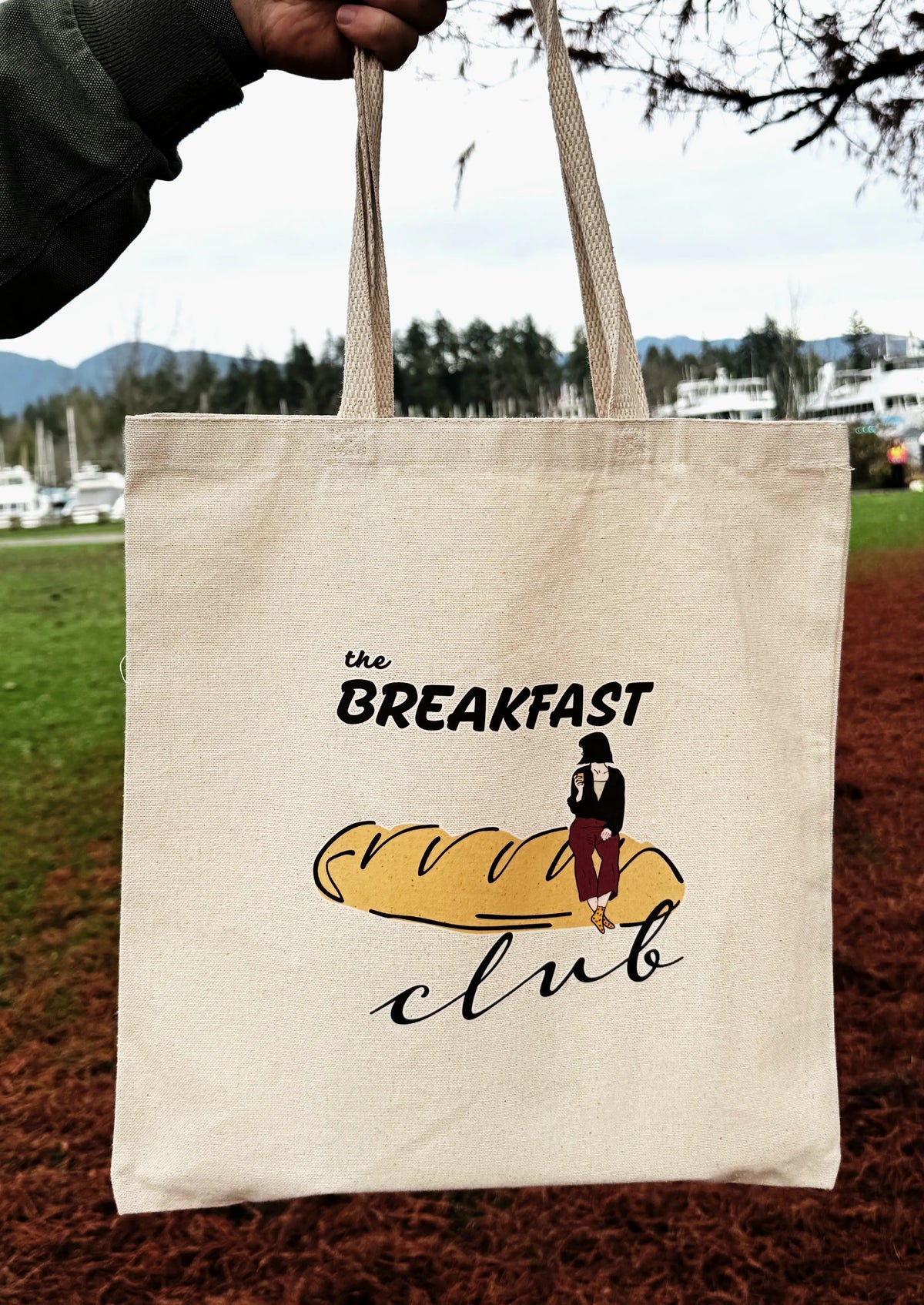 Breakfast Club canvas tote bag worn over the shoulder, featuring line art bread illustration, photographed outdoors.