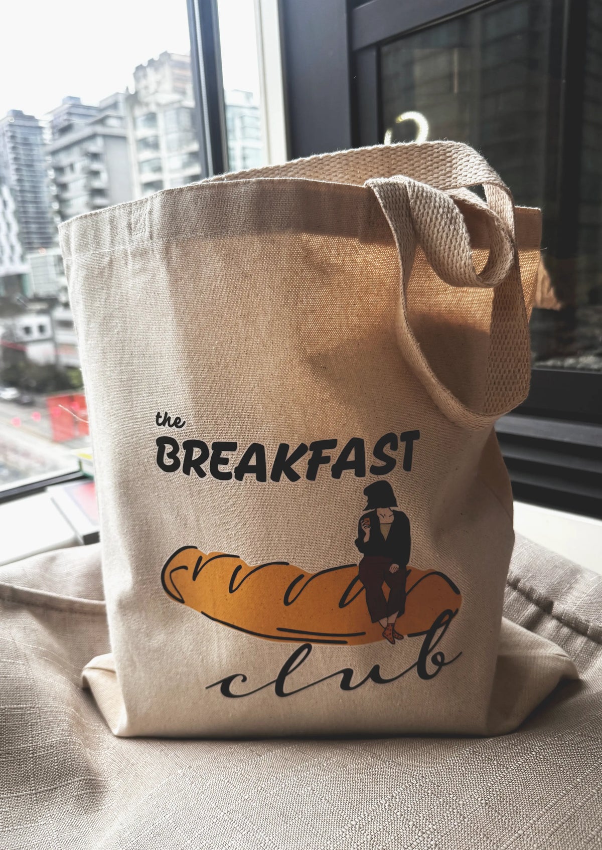 Breakfast Club canvas tote bag standing upright on a table near a window with city buildings in the background.