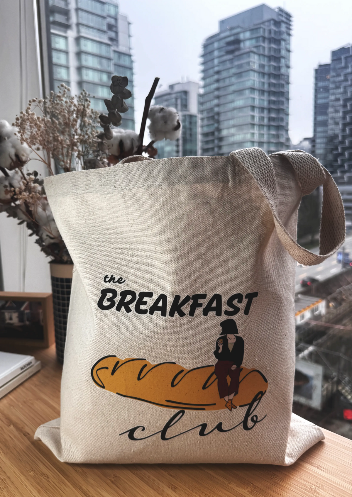 Side view of Breakfast Club canvas tote bag with bread illustration, photographed indoors near a window.