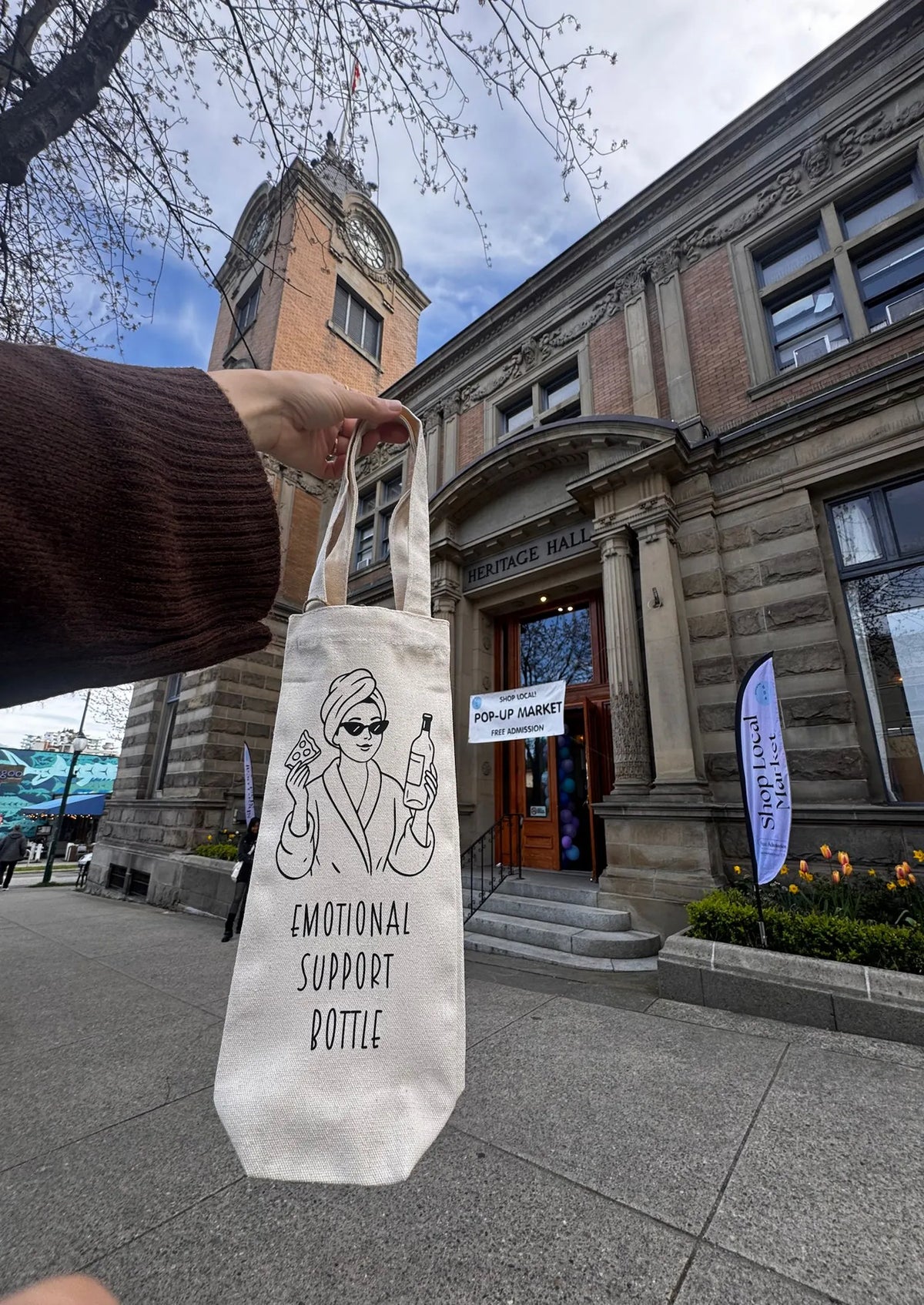 Person holding a tote bag with 'Emotional Support Bottle' design in front of a building.