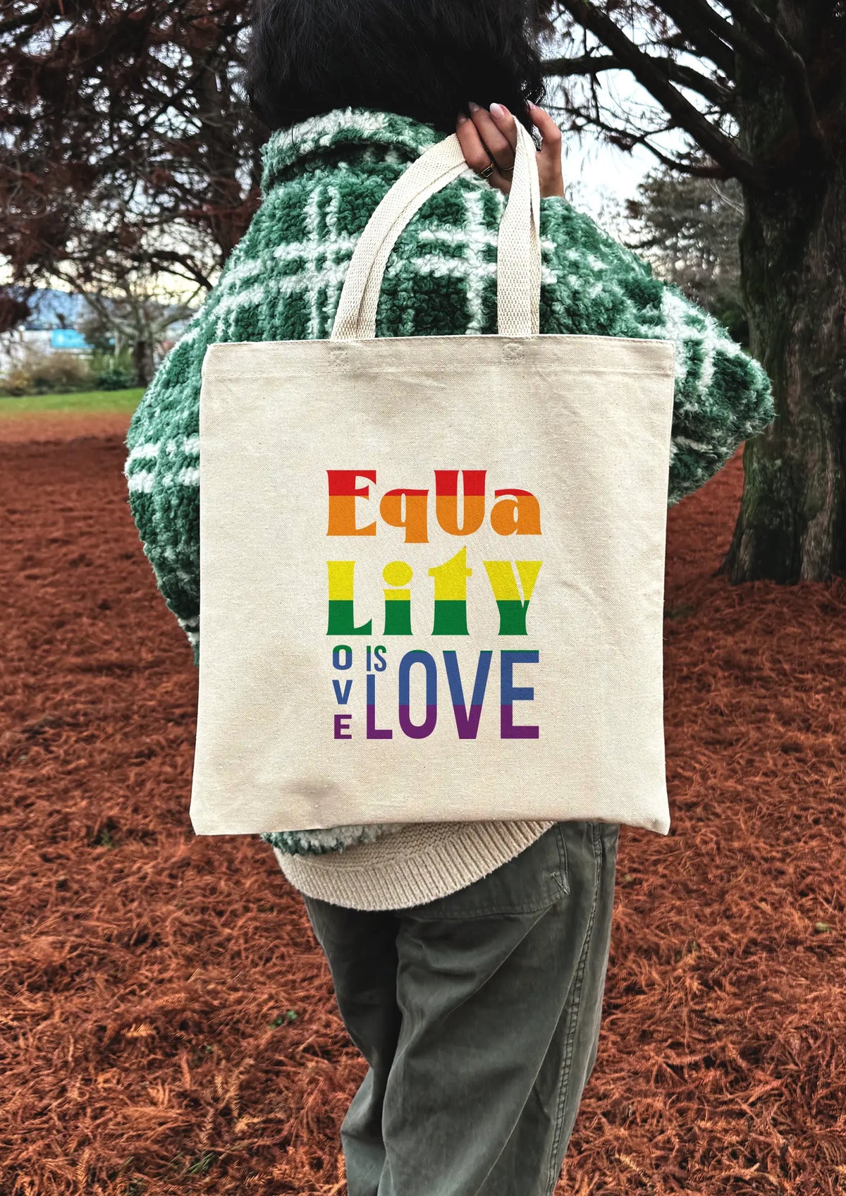 Person holding a tote bag with 'Equality is Love' text in rainbow colors outdoors.