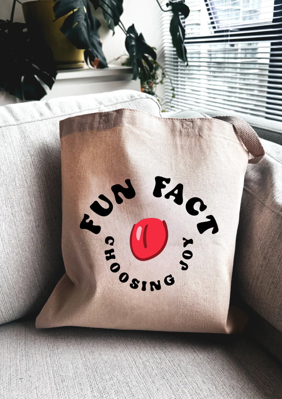 Beige tote bag with 'FUN FACT CHOOSING' text and red circle on a gray couch.