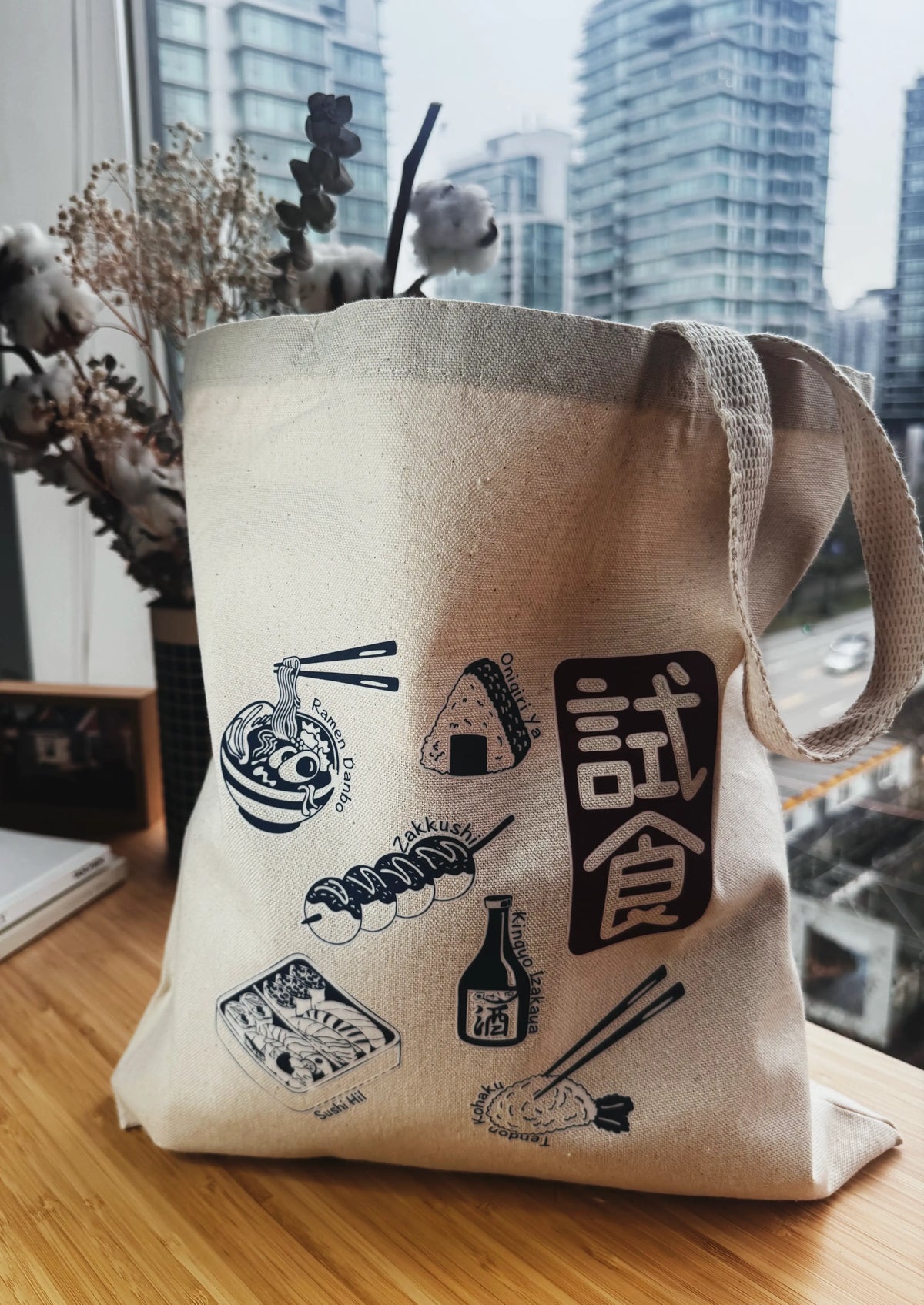 Beige tote bag with black illustrations and text on a wooden surface with a cityscape background