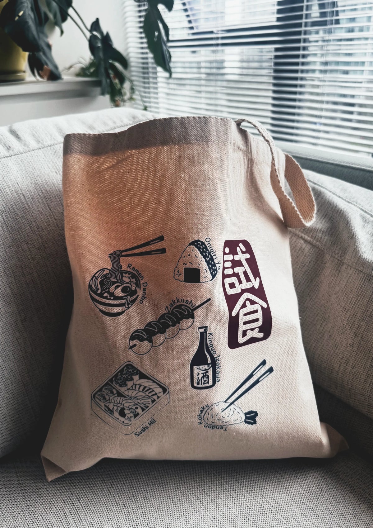 Beige tote bag with Japanese food illustrations and text on a couch.
