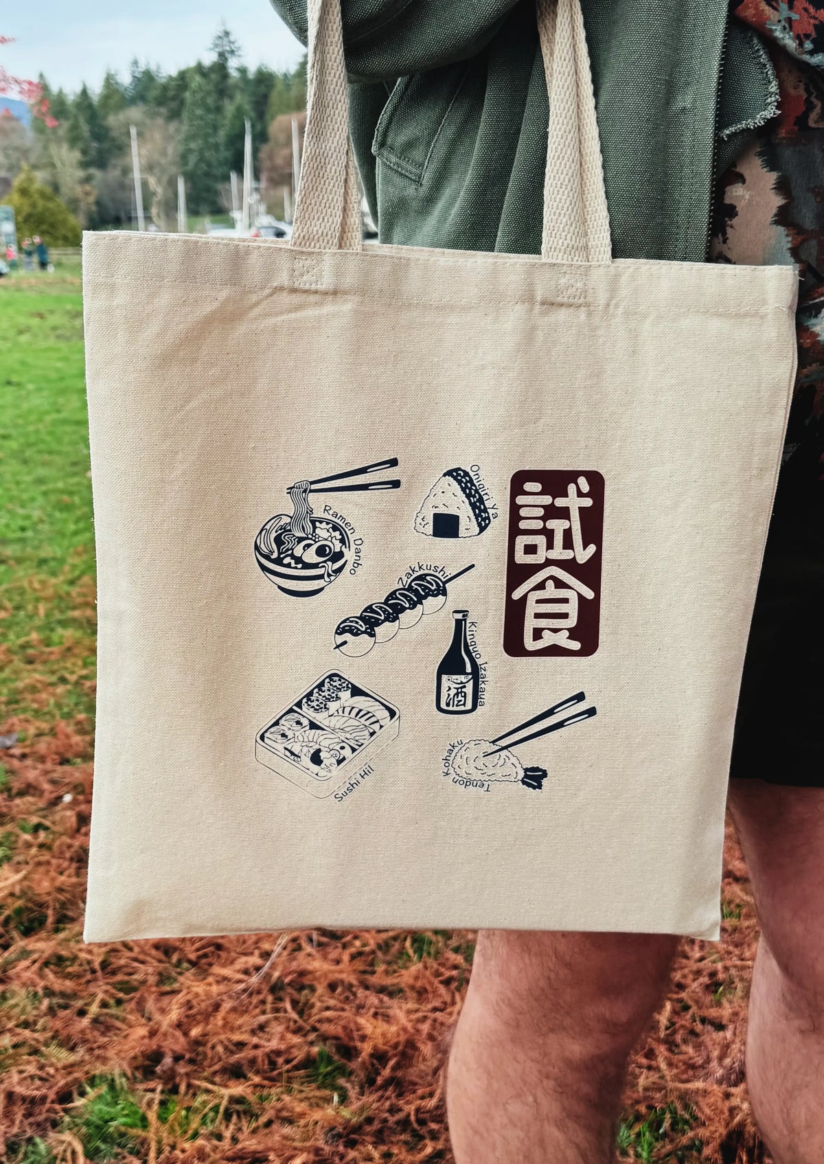Beige tote bag with Japanese food illustrations and text held by a person outdoors.