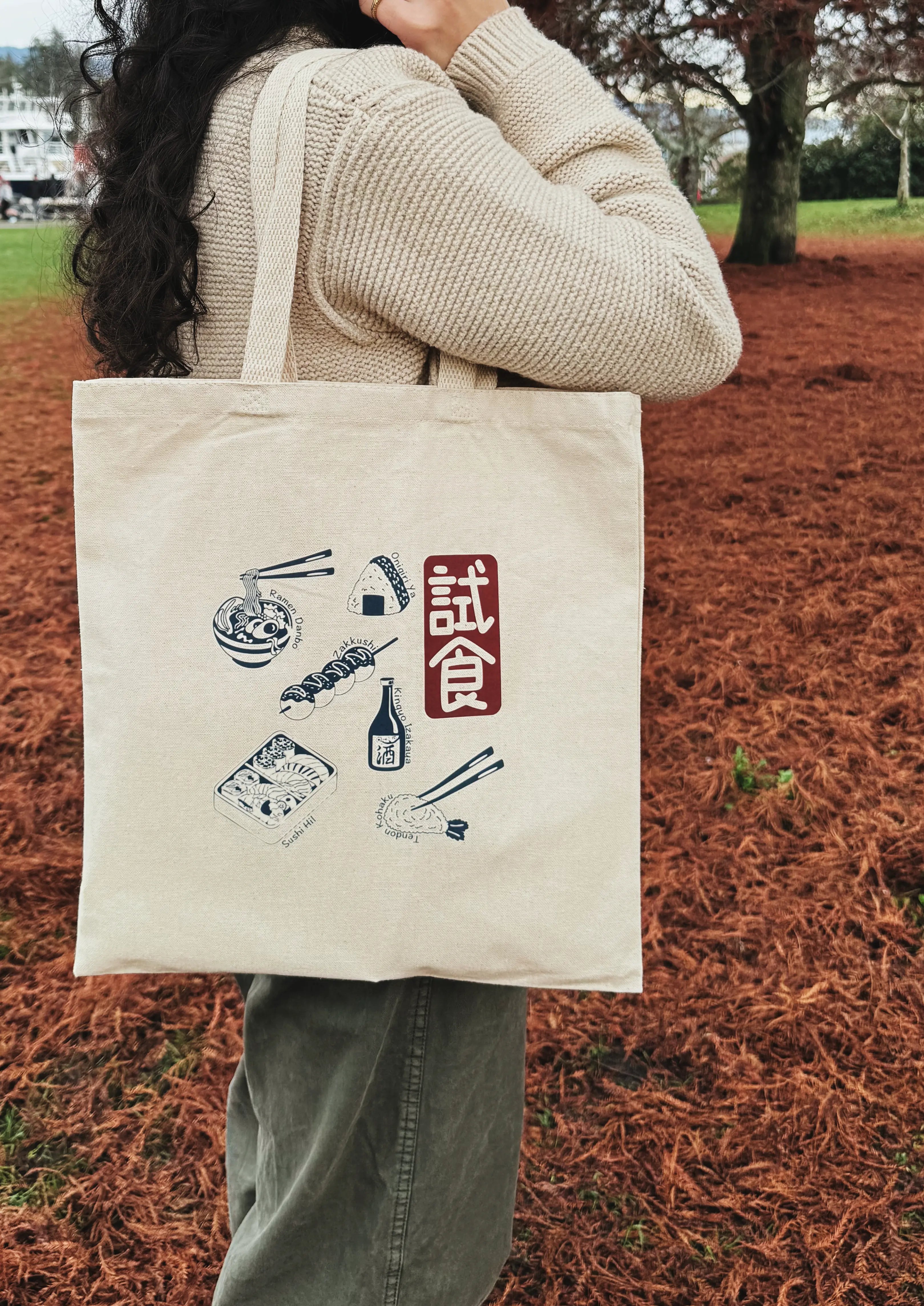 Person holding a tote bag with Japanese food illustrations and text outdoors.