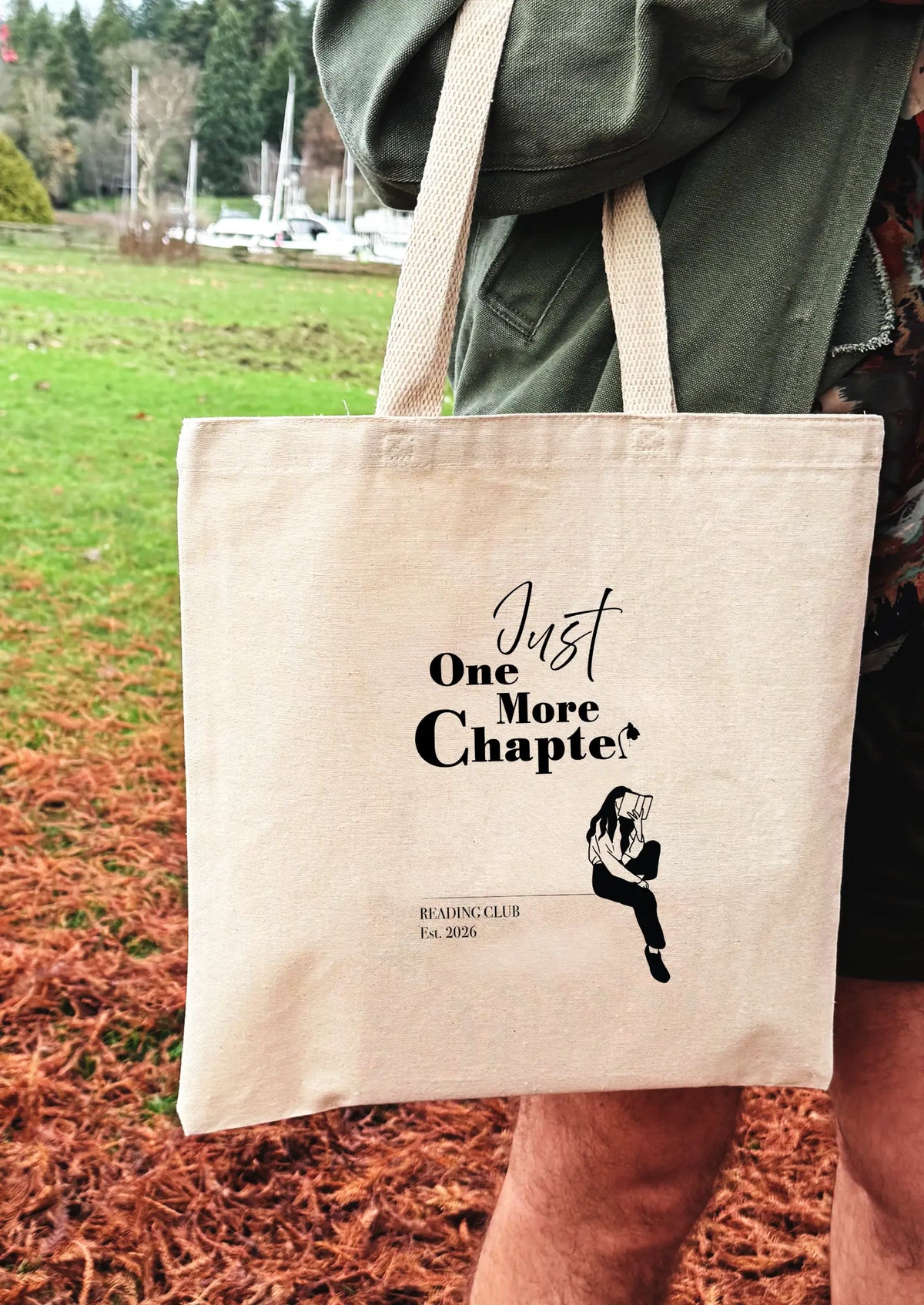 Tote bag with 'Just One More Chapter' text and reading club design held by a person outdoors.