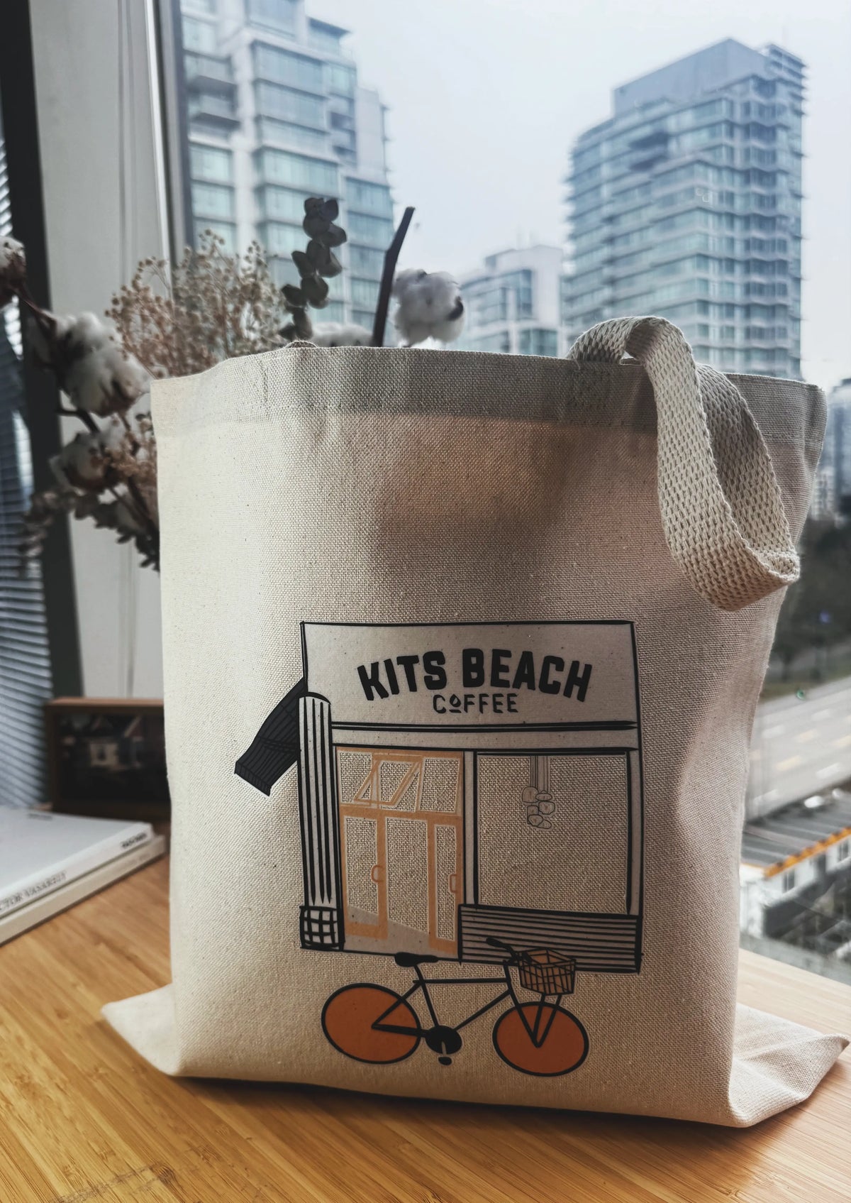 Tote bag with 'Kits Beach Coffee' design on a wooden surface with a cityscape background