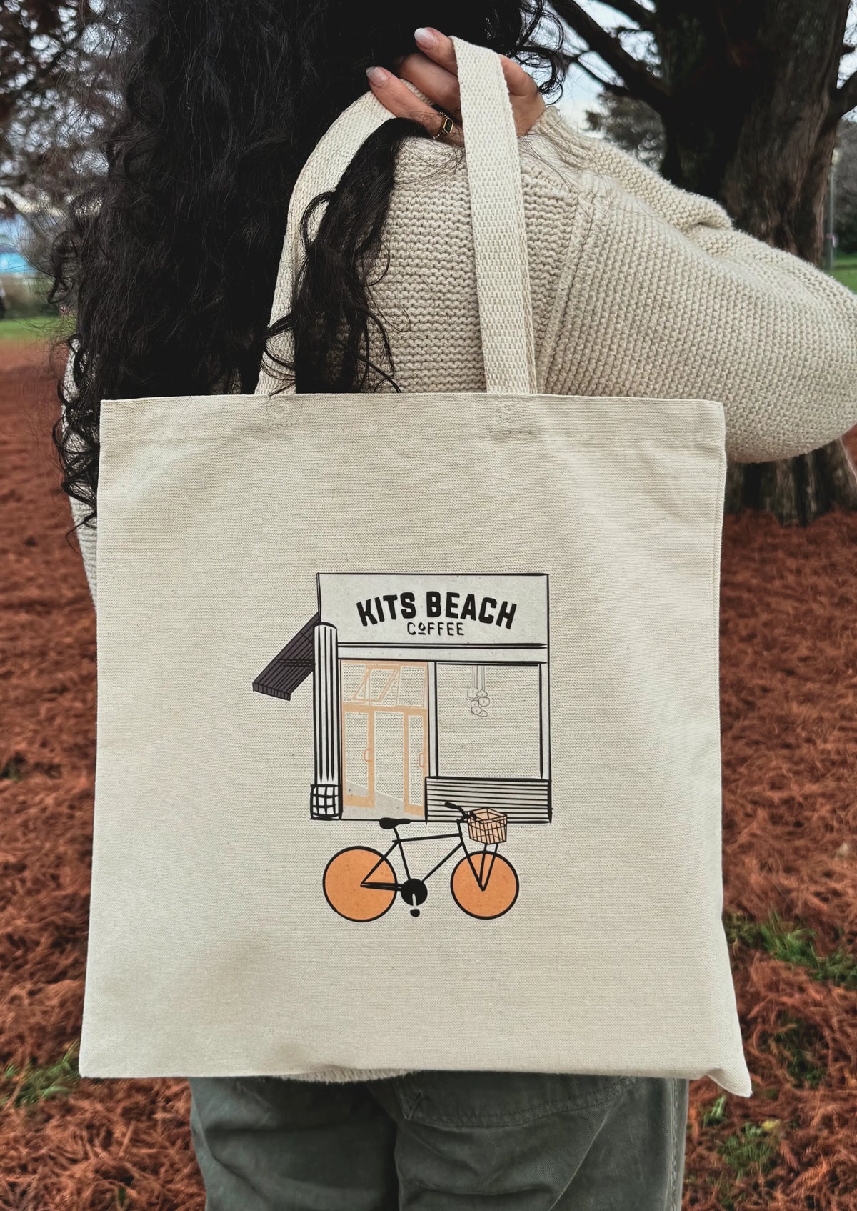 Person holding a tote bag with 'Kits Beach Coffee' design outdoors.