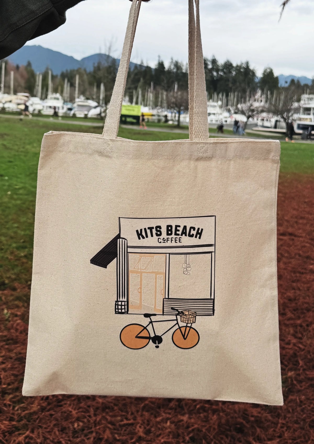 Beige tote bag with 'Kits Beach Coffee' graphic on a grassy field background