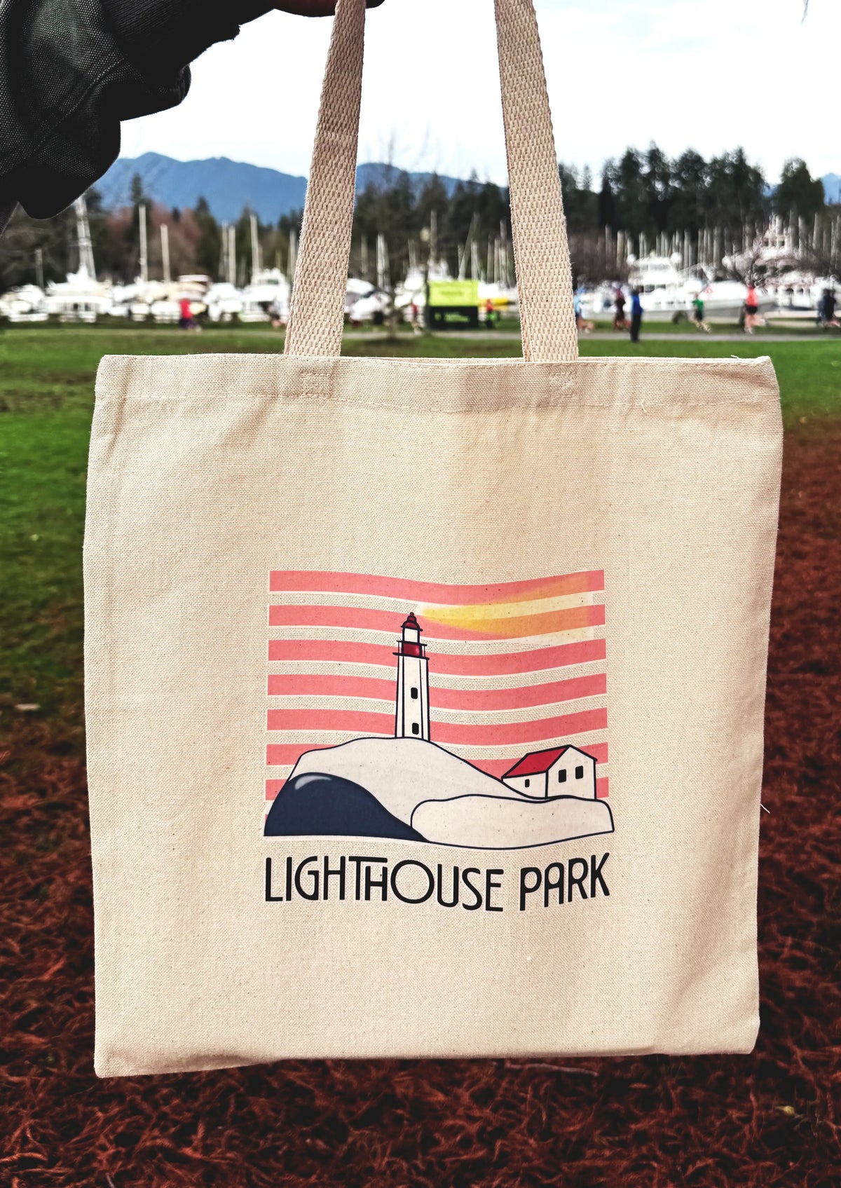 Tote bag with 'Lighthouse Park' design held outdoors with a scenic background.