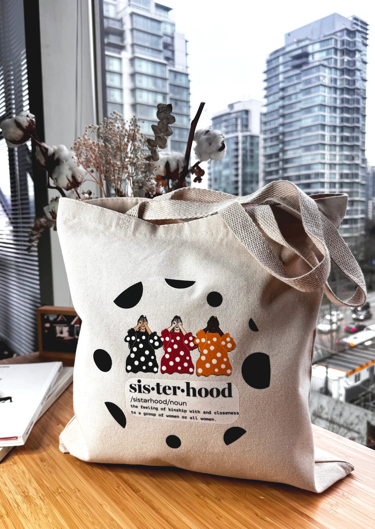 Sisterhood line art canvas tote bag displayed indoors on a wooden table with city skyline and dried florals