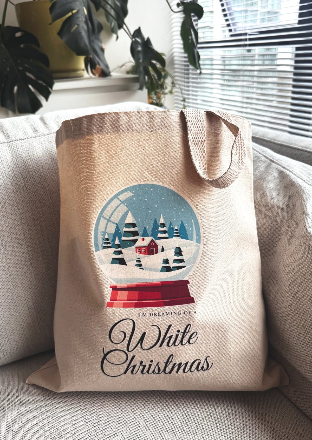 Snow globe winter scene canvas tote bag displayed indoors on a couch with houseplant and window light