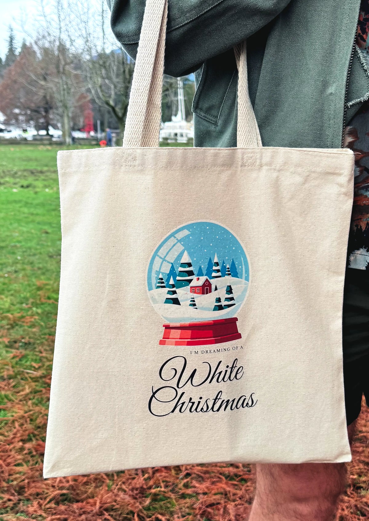 Front view of snow globe winter scene canvas tote bag being held outdoors with winter park setting