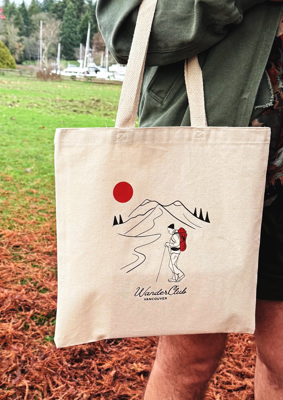 Tote bag with a design of a person hiking with mountains and a red sun, held by a person outdoors.
