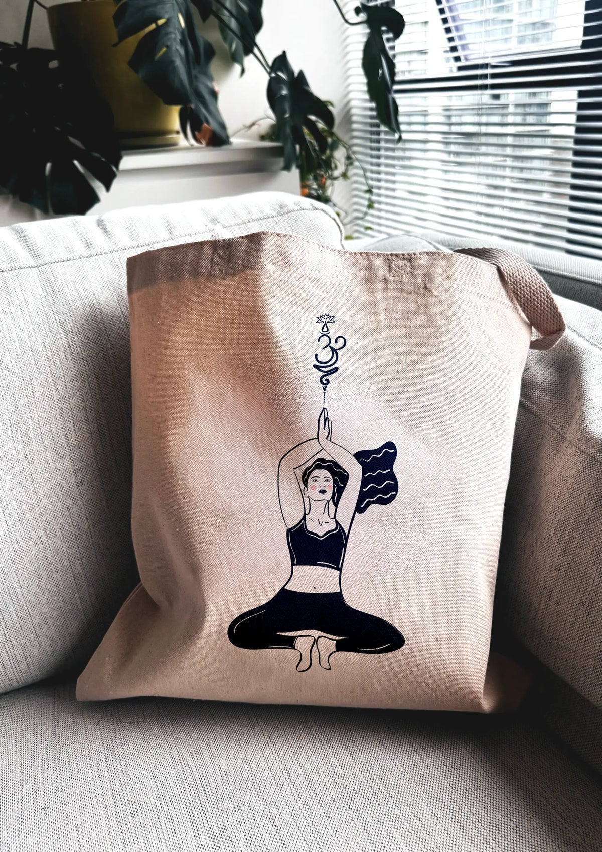 Yoga Flow canvas tote bag with line art illustration of a woman in a seated yoga pose, photographed indoors on a couch near a window.