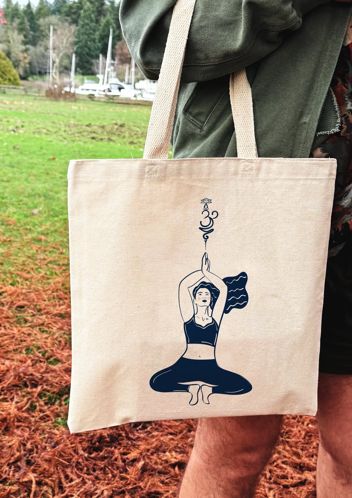 Yoga Flow canvas tote bag featuring a seated yoga illustration, held outdoors in a park setting.