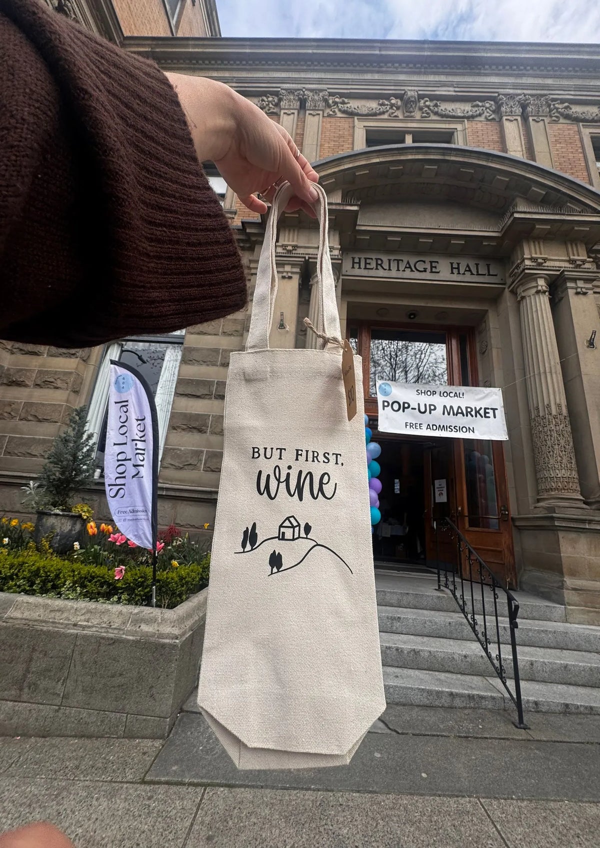 Person holding a tote bag with 'But first, wine' text in front of Heritage Hall.