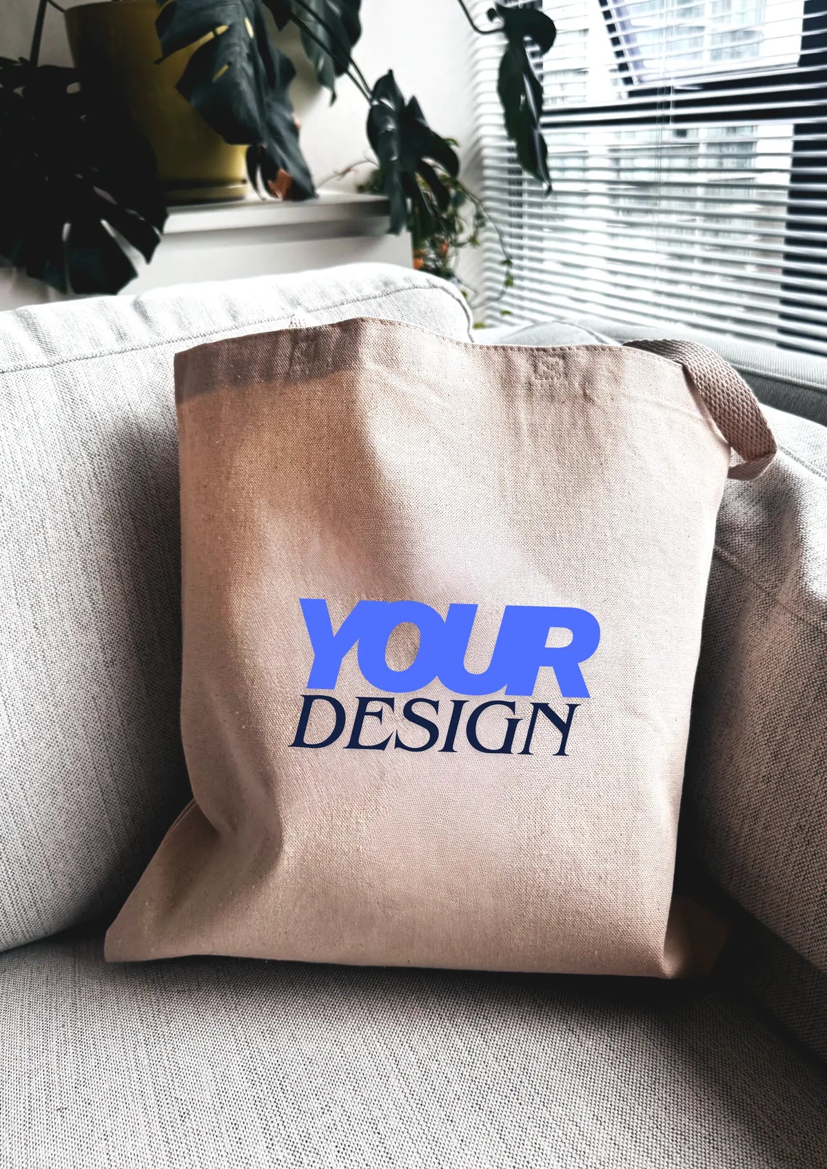 Beige tote bag with 'YOUR DESIGN' text on a gray couch.