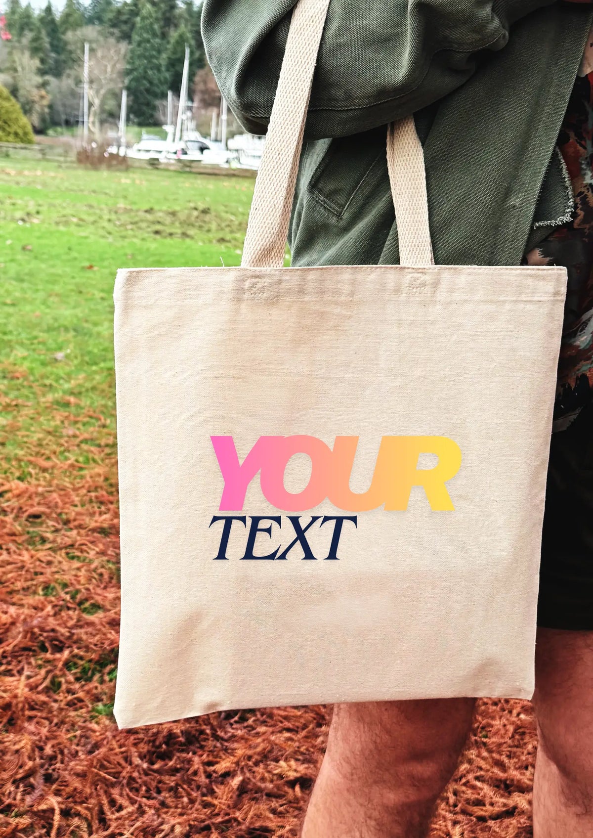 Beige tote bag with 'YOUR TEXT' in colorful letters held by a person outdoors.