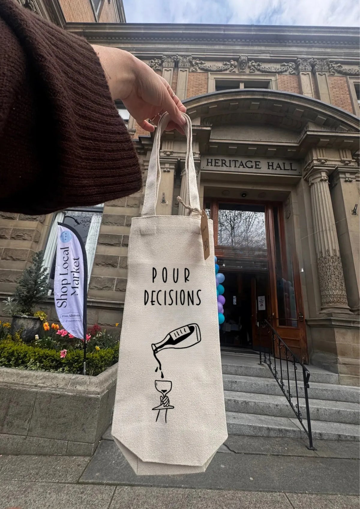 Person holding a tote bag with 'Pour Decisions' text and bottle graphic in front of Heritage Hall.