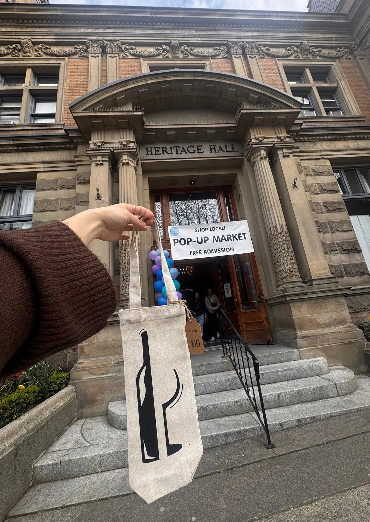 Person holding a tote bag with a bottle design in front of Heritage Hall with a pop-up market sign.
