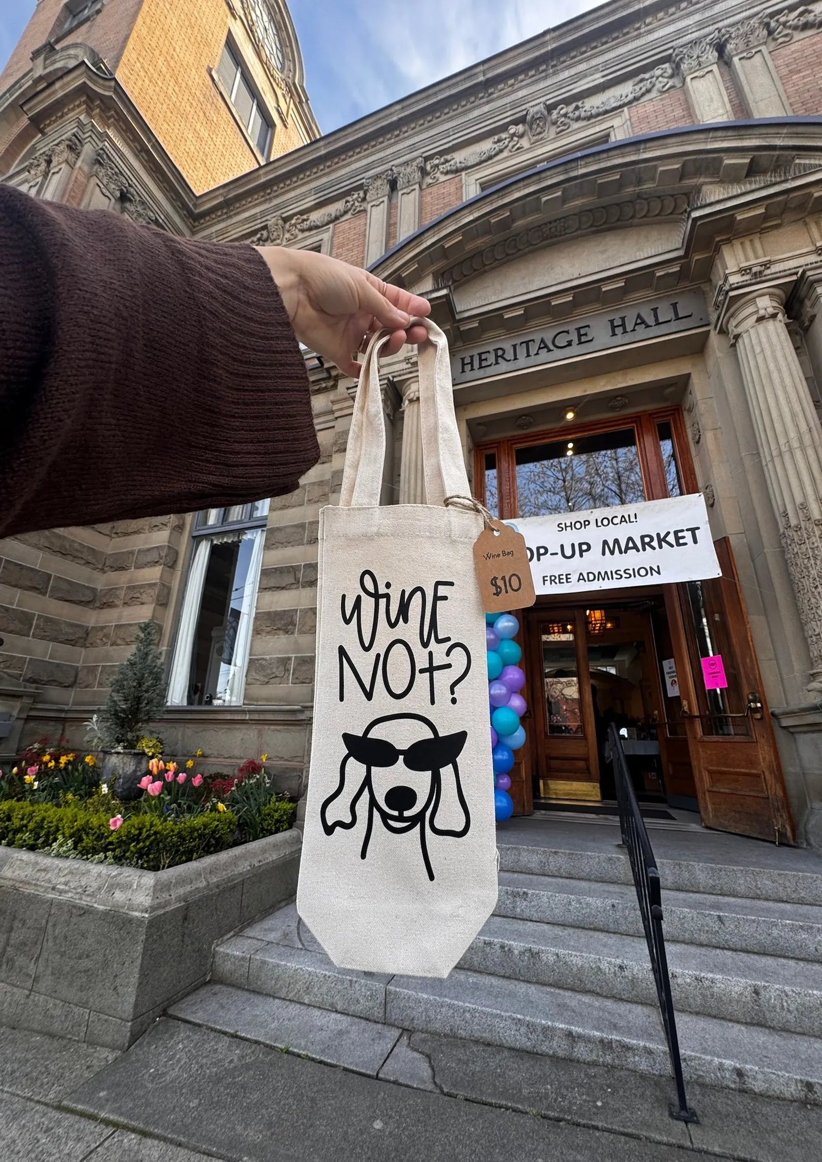 Person holding a tote bag with 'Wine Not?' design in front of Heritage Hall.